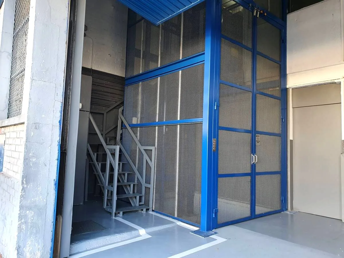 Montacargas industrial con hueco cerrado de malla y escalera de acceso, instalado en la fachada de una nave