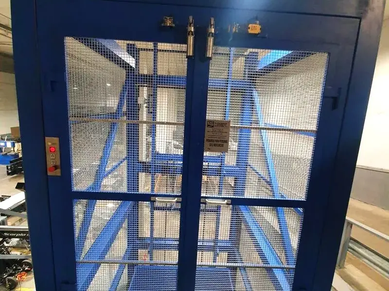 Montacargas de malla azul con puertas de dos hojas y su botonera, en una planta de nave