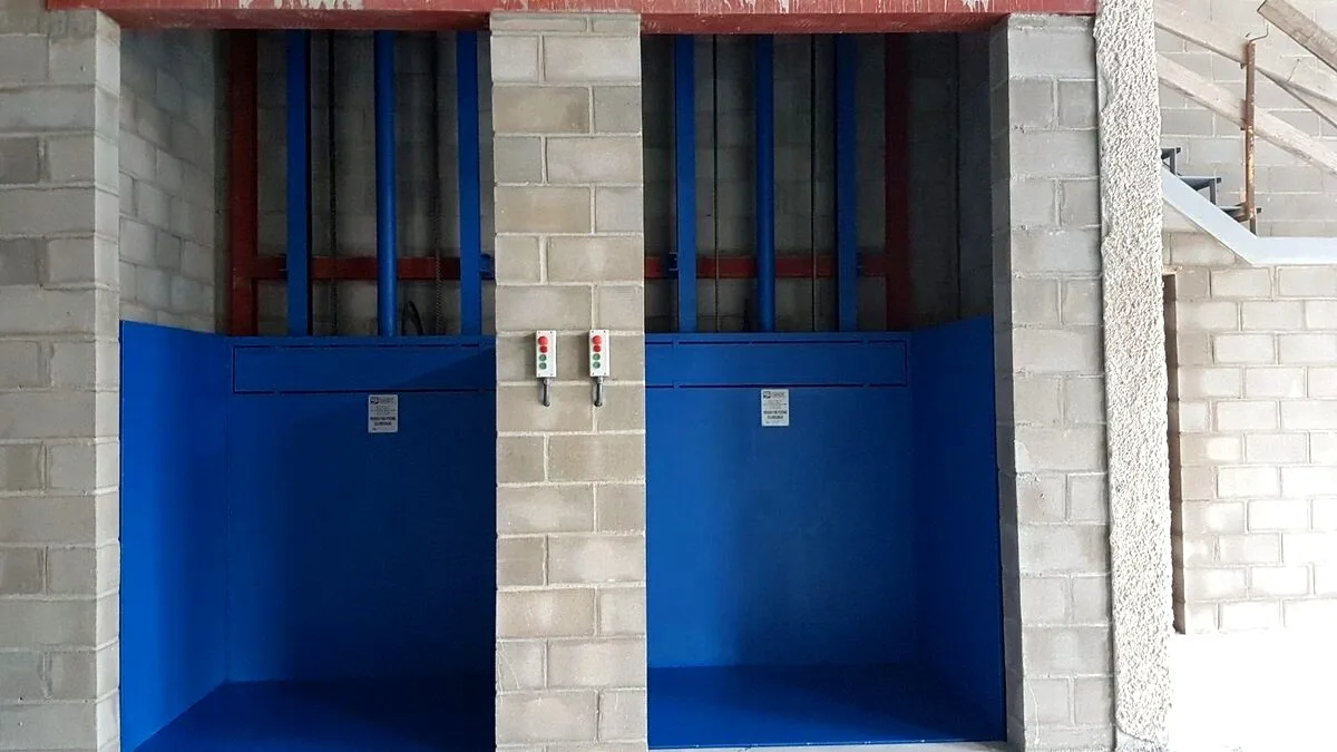 Dos montacargas de cabina azul instalados uno junto al otro, con sus botoneras de mando en el pilar central
